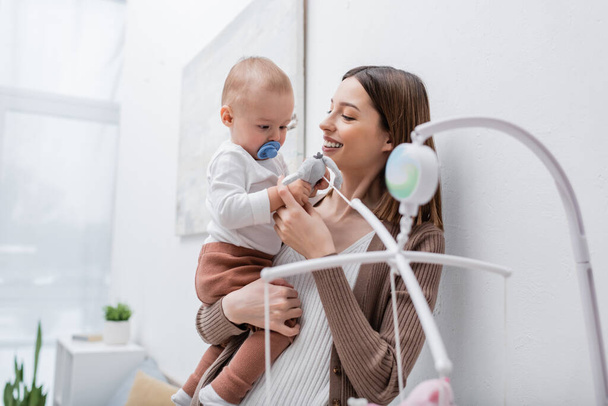 Smiling mother holding baby boy near toys at home  - Photo, Image