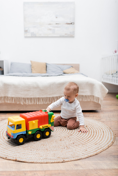 Mały chłopiec ze smoczkiem bawiący się zabawką na podłodze w sypialni  - Zdjęcie, obraz