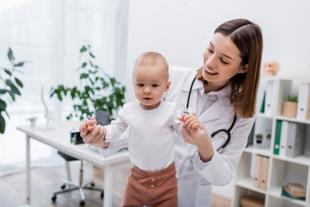 Семейный врач в белом халате держит за руки младенца в клинике - Фото, изображение