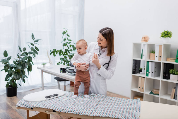 Medico di famiglia che tiene il bambino vicino alla scala elettronica sul divano medico in clinica  - Foto, immagini