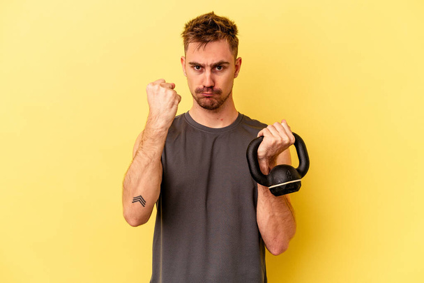 Молодой спортсмен держит гантель на желтом фоне, показывая кулак к камере, агрессивное выражение лица. - Фото, изображение