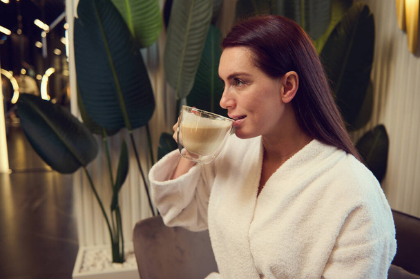 Side portrait of elegant beautiful dark haired middle aged woman in white terry bathrobe drinking cappuccino during coffee break relaxing in spa after professional beauty therapy. Cosmetology concept - Φωτογραφία, εικόνα