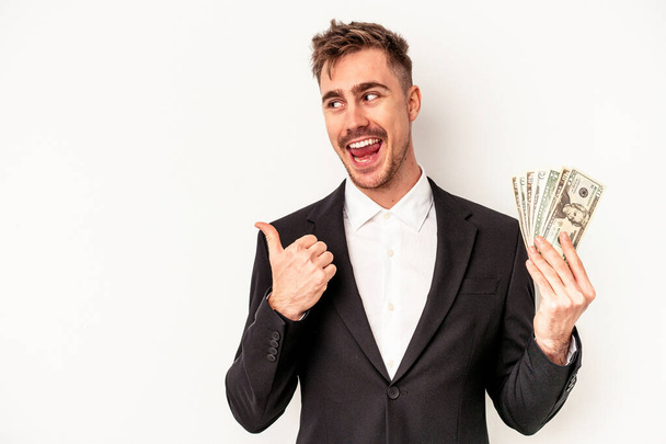 Joven hombre de negocios caucásico sosteniendo billetes de banco aislados en puntos de fondo blanco con el dedo pulgar lejos, riendo y sin preocupaciones. - Foto, imagen