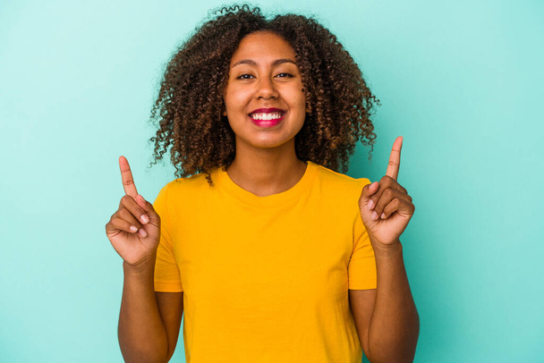 Giovane donna afroamericana con i capelli ricci isolati su sfondo blu indica con entrambe le dita anteriori in alto mostrando uno spazio vuoto. - Foto, immagini