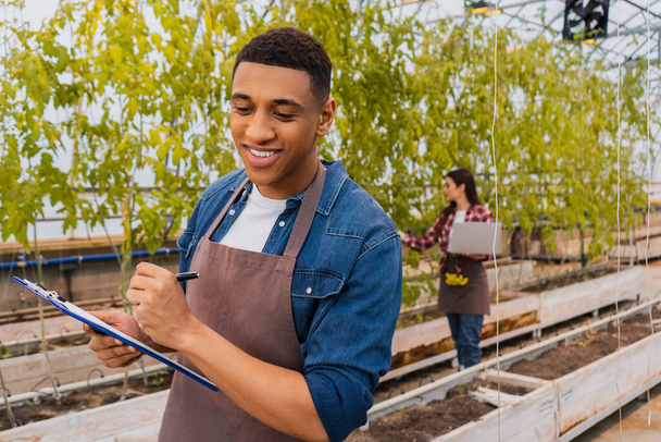 Sonriente agricultor afroamericano sujetando portapapeles cerca de colega borrosa con portátil en invernadero  - Foto, Imagen
