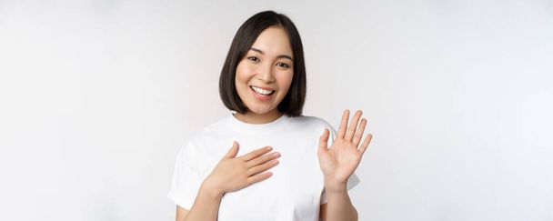 Retrato de hermosa chica coreana levantando la mano, presentarse, poner la mano en el corazón, saludo, de pie sobre fondo blanco - Foto, imagen