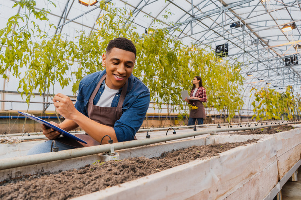 Happy african american farmer holding clipboard near ground in greenhouse  - Photo, Image