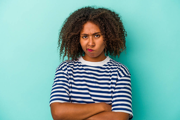Jonge Afro-Amerikaanse vrouw met krullend haar geïsoleerd op blauwe achtergrond fronsend gezicht in ongenoegen, houdt armen gevouwen. - Foto, afbeelding