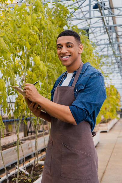 Positive african american farmer in apron writing on clipboard near plants in greenhouse  - Foto, afbeelding