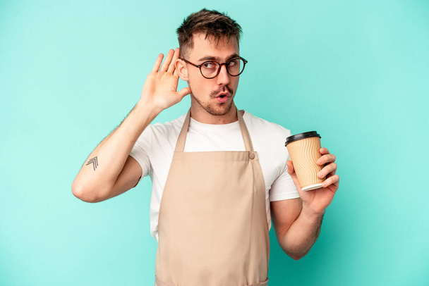 Young store clerk man holding a coffee isolated on blue background trying to listening a gossip. - Fotó, kép
