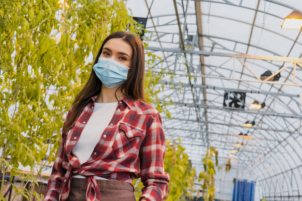Farmer in medical mask looking at camera near plants in greenhouse  - Photo, Image
