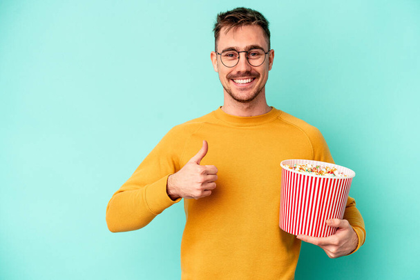 Junger kaukasischer Mann isst Popcorn isoliert auf blauem Hintergrund, lächelt und hebt den Daumen - Foto, Bild