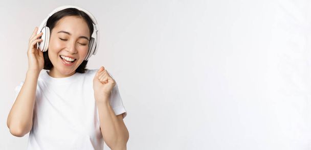 Σύγχρονη ασιατική κοπέλα χορεύει, ακούει μουσική με ακουστικά, χαμογελά ευτυχισμένη, στέκεται σε μπλουζάκι πάνω από λευκό φόντο - Φωτογραφία, εικόνα