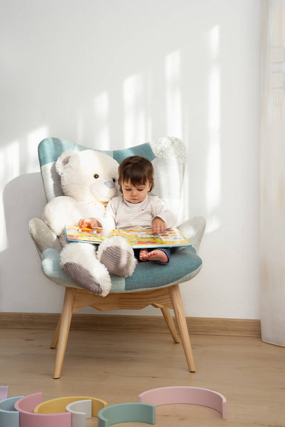 Pieni tyttö, jolla on keskittynyt ele, istuu nallekarhun kanssa nojatuolilla, kun hän etsii satukirjaa huoneessaan aurinkoisena aamuna..  - Valokuva, kuva