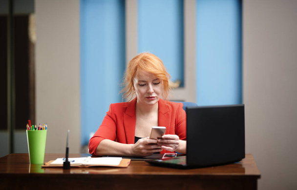 Ofiste dizüstü bilgisayar ve akıllı telefon kullanan kızıl saçlı bir iş kadını. - Fotoğraf, Görsel