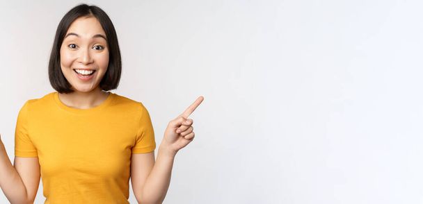 Cute asian girl pointing fingers sideways, showing left and right promo, two choices, variants of products, standing in yellow tshirt over white background - Photo, Image