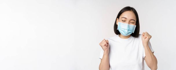 Covid-19, healthcare and medical concept. Enthusiastic asian woman in medical face mask, dancing and celebrating, winning, achieve goal, standing over white background - Фото, изображение