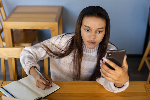 ビジネス、テクノロジー、ライフスタイル、人々の概念-コーヒーショップでのオンライン会議中にノートブックにペンで重要な情報を書いて電話で深刻な暗い髪の女の子 - 写真・画像