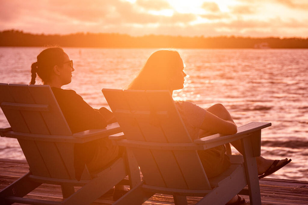 Two young women sitting at the beach and enjoying the sunset over the water - Photo, image