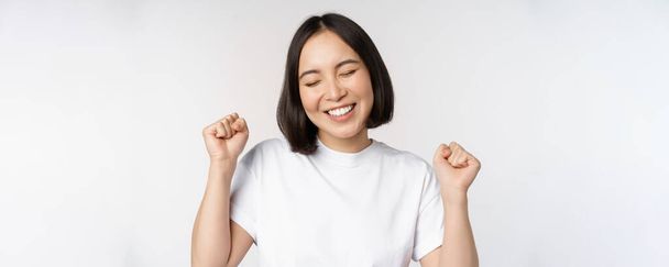 Dancing asian girl celebrating, feeling happy and upbeat, smiling broadly, standing over studio white background - Photo, Image