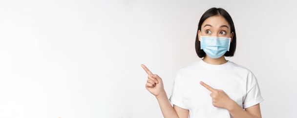 Young korean woman in medical face mask pointing fingers left and looking at logo, showing advertisement or banner, standing over white background - Valokuva, kuva