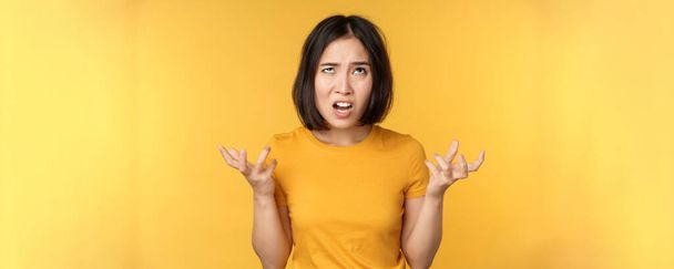 Immagine di donna asiatica arrabbiata, urlando e maledicendo, guardando indignata, furiosa espressione del viso, in piedi su sfondo giallo - Foto, immagini