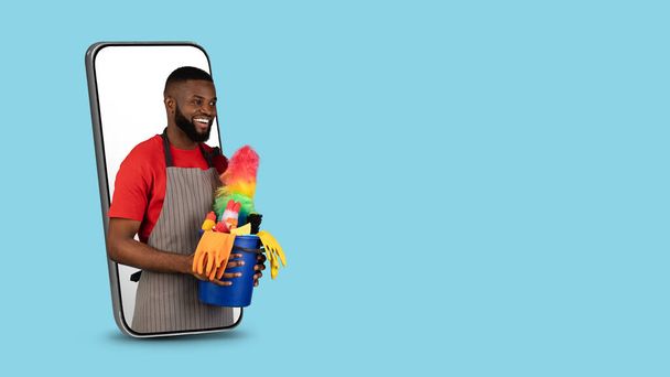 Limpiador masculino negro con cesta con detergentes asomándose a la pantalla del teléfono inteligente - Foto, imagen