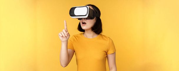 Lány a VR-ben. Gyönyörű fiatal ázsiai nő, a virtuális valóság szemüveg és a játék, csevegés virtuálisan, álló sárga háttér - Fotó, kép