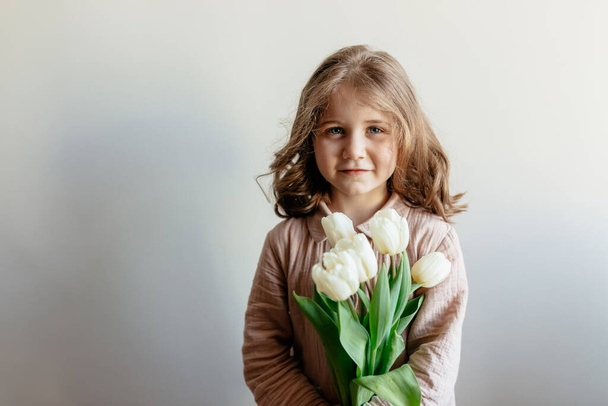Portrait of charming little girl with wavy hairstyle wearing home suit is holding white flowers over isolated background. Indoor photo of happy little girl is celebrating her birthday.  - Foto, immagini