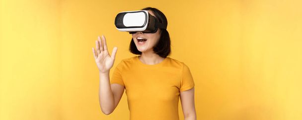 Chica en VR. Hermosa joven mujer asiática, usando gafas de realidad virtual y jugando, charlando virtualmente, de pie sobre fondo amarillo - Foto, Imagen