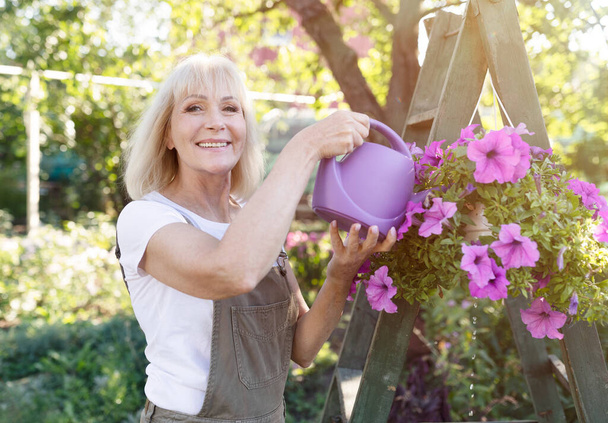 Ocio, concepto de estilo de vida. Mujer madura feliz regando flores de petunia en macetas, jardinería en su propio jardín de verano - Foto, imagen