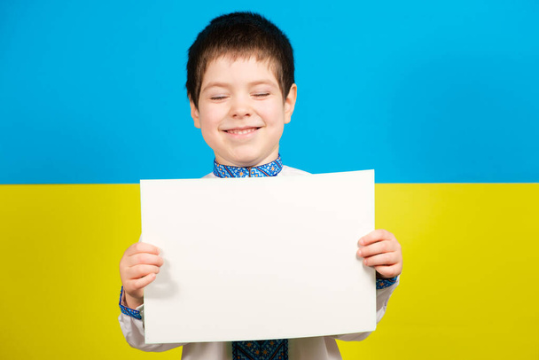 A happy child in a Ukrainian national embroidered shirt holds a blank sheet for text against the background of a yellow blue flag - Photo, Image