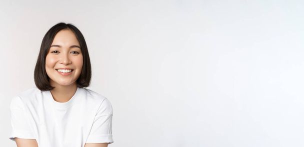 Hermosa chica coreana sonriendo, dientes blancos, con un aspecto encantador en la cámara, de pie en camiseta blanca sobre fondo del estudio - Foto, imagen