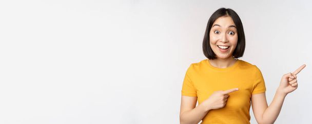Hämmästynyt nuori aasialainen nainen, joka näyttää mainoksen syrjään, osoittaa sormella suoraan myynninedistämistekstiin, tuotemerkkiin, seisoo onnellisena valkoista taustaa vasten - Valokuva, kuva