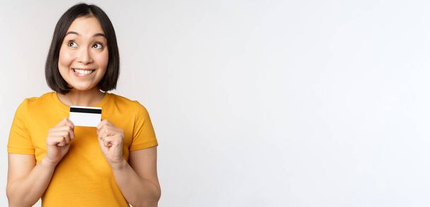 Portrait of beautiful korean girl holding credit card, recommending bank service, standing in yellow tshirt over white background - Foto, Imagem