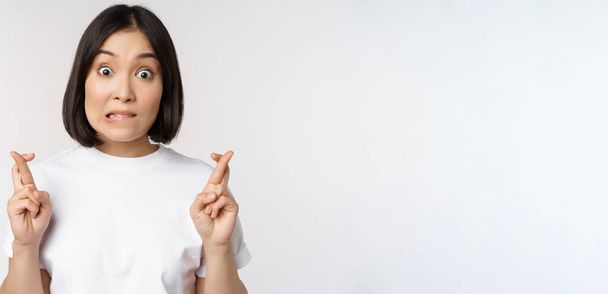 esperançoso asiático menina cruz dedos, fazendo desejo, antecipando, desejando smth, de pé em tshirt sobre branco fundo - Foto, Imagem