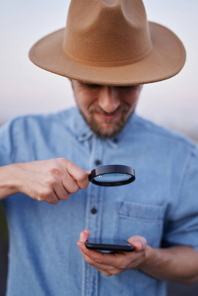 Young man in hat looking with magnifying glass at smartphone - Photo, Image