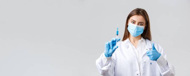 Covid-19, virüsü önleme, sağlık çalışanları ve karantina konsepti. Tıbbi maske ve lastik eldiven takmış kararlı genç doktor, önlükler baş parmak ve şırıngada koronavirüs aşısı gösteriyor. - Fotoğraf, Görsel
