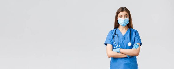 Covid-19, preventing virus, health, healthcare workers and quarantine concept. Amused female doctor or nurse in blue scrubs with stethoscope and personal protective equipment look intrigued - Фото, зображення