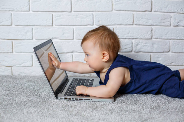 Mavi pamuklu tulumlu 6-7 aylık küçük bir çocuk açık bir dizüstü bilgisayarla oturur ve eğitici oyunlar izler. - Fotoğraf, Görsel