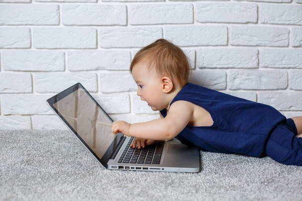 Mavi pamuklu tulumlu 6-7 aylık küçük bir çocuk açık bir dizüstü bilgisayarla oturur ve eğitici oyunlar izler. - Fotoğraf, Görsel