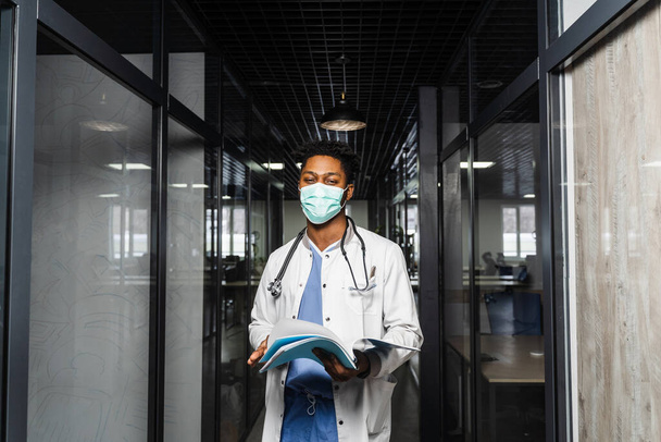Tıbbi klinikte kitapları olan maskeli Afrikalı bir doktor. Siyahi tıp öğrencisi. - Fotoğraf, Görsel