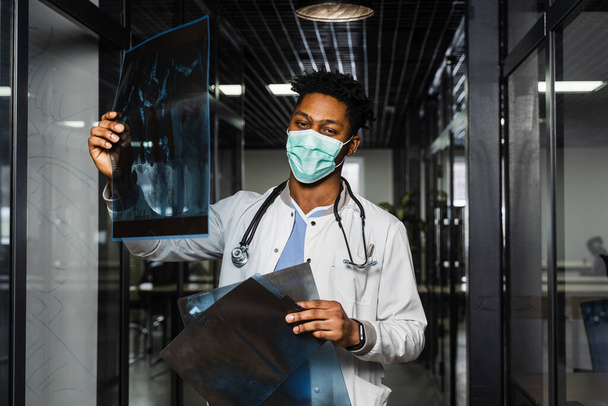 Αφρικανός γιατρός εξετάζει τις ακτινογραφίες σε μια ιατρική κλινική. Μαύρος φοιτητής με ιατρική μάσκα μελετά και εξετάζει αξονική τομογραφία - Φωτογραφία, εικόνα