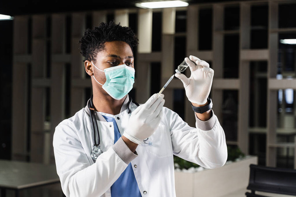 Αφρικανός γιατρός με ιατρική μάσκα ετοιμάζεται να κάνει ένεση με το εμβόλιο Coronavirus covid-19. Μαύρος γιατρός με λευκή ιατρική ρόμπα με σύριγγα για εμβολιασμό - Φωτογραφία, εικόνα