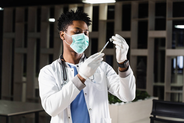 Afrikkalainen lääkäri lääketieteellisessä naamiossa valmistautuu pistämään sepelvaltimoviruksen covid-19-rokotteen. Musta lääkäri valkoisessa kylpytakissa ruiskulla rokotusta varten - Valokuva, kuva