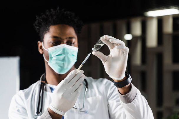 Afrikkalainen lääkäri lääketieteellisessä naamiossa valmistautuu pistämään sepelvaltimoviruksen covid-19-rokotteen. Musta lääkäri valkoisessa kylpytakissa ruiskulla rokotusta varten - Valokuva, kuva