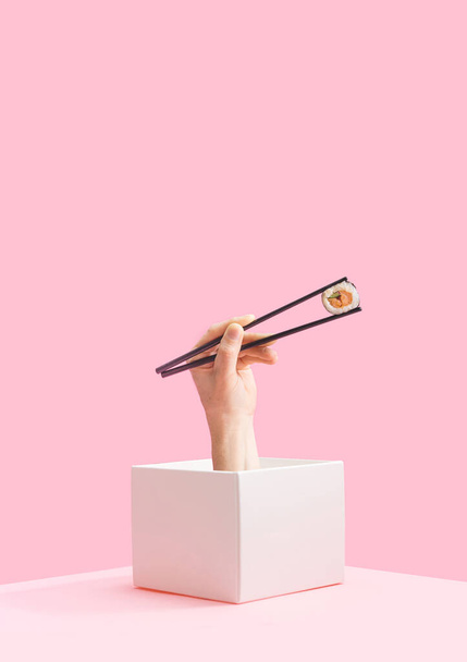 Минимальная концепция суши на вынос. Мужская рука с черной палочкой выскакивает из коробки с одним кусочком рисового лосося и огуречным рулетом из сушеных морских водорослей. Детский розовый фон - Фото, изображение