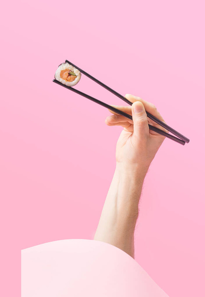 Sushi takeaway minimal concept. Man hand holding black chopstick pop up from round box with one piece rice salmon and cucumber roll in nori dried edible seaweed. Awesome pink background - Photo, Image