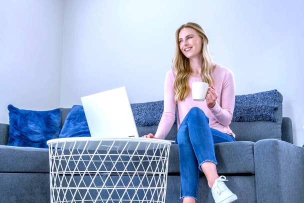 Sarışın, güzel bir kadın dizüstü bilgisayarda çalışıyor ve evdeki kanepede oturuyor.  - Fotoğraf, Görsel