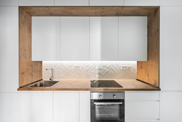 Beyaz dolaplı mutfak ve tezgahın yanında ahşap çerçeveli çelik lavabo ve fırın. - Fotoğraf, Görsel